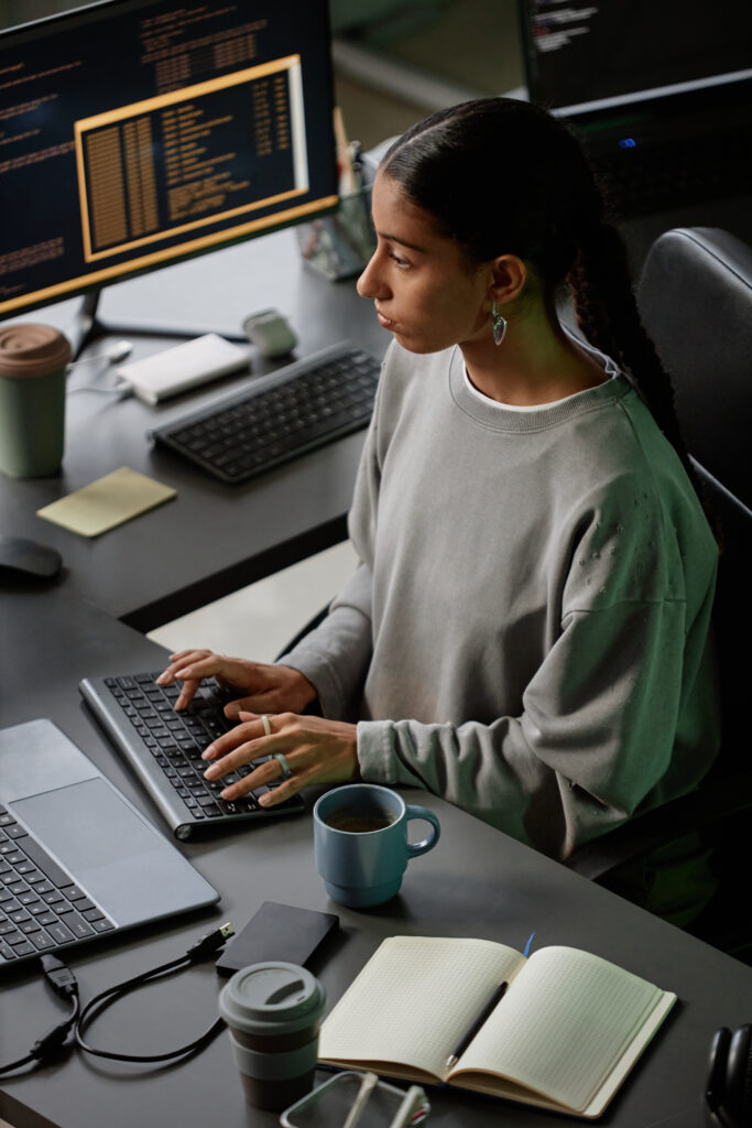 High angle of young Middle Eastern female programmer immersed in coding on keyboard of desktop PC, while working at cluttered with stationery and cups desk in office